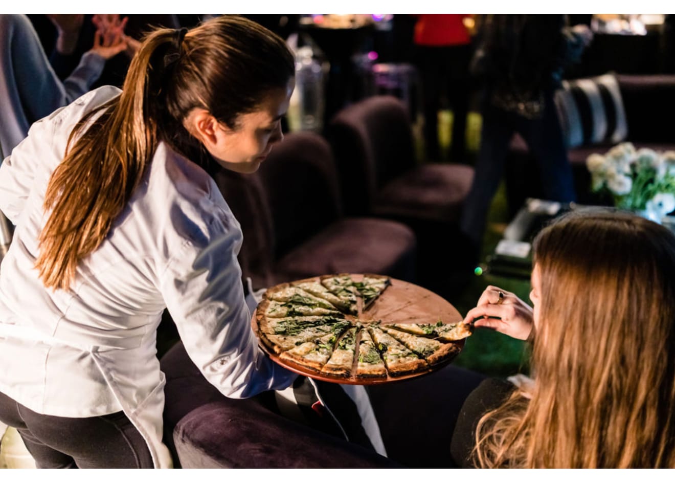 Server presenting a sliced white pizza to a seated guest at an upscale evening event in Los Angeles